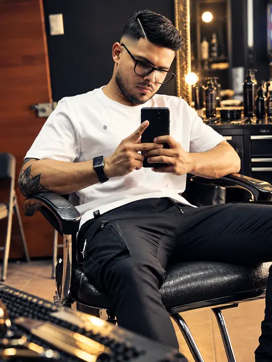 Steven Baltodano sentado en la silla de barbero de su local, revisando el celular